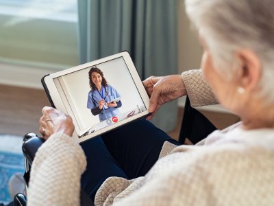Back view of senior woman sitting in wheelchair making video call with her doctor while staying at home during covid pandemic. Close up of helpful general practitioner in video conference with old woman on digital tablet. Sick woman in online consultation from home: distance and telehealth concept.